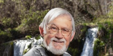 Older man with glasses smiling outdoors near a waterfall.