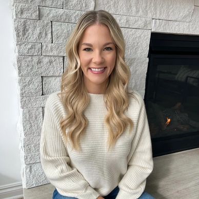 Smiling woman with blonde hair sitting by a fireplace.