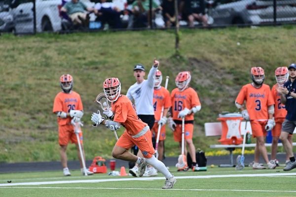 Lacrosse player running during a game, teammates and coach watching.