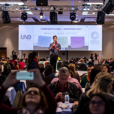 Reinaldo no palco com microfone e telão atrás dando palestra para 500 professores.