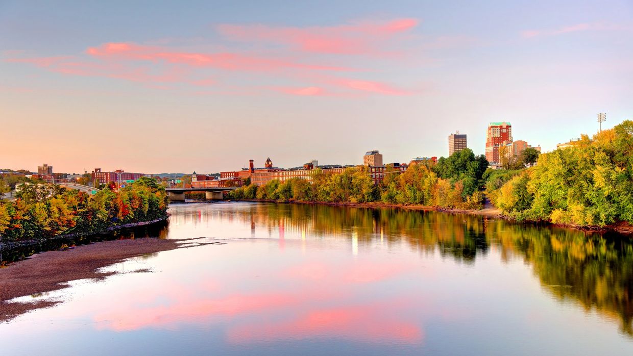 City skyline reflected in a calm river at sunset with autumn trees.