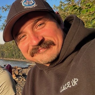 Man with a mustache smiling outdoors near a lake in a hoodie and cap.