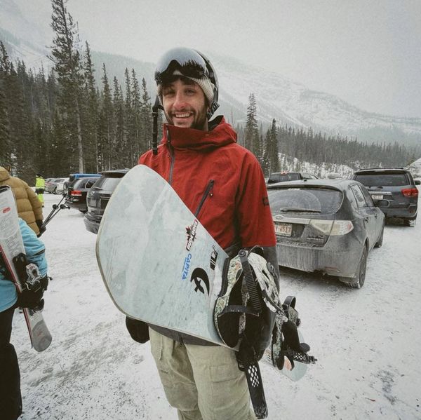 Smiling snowboarder in red jacket holding snowboard in snowy parking lot.