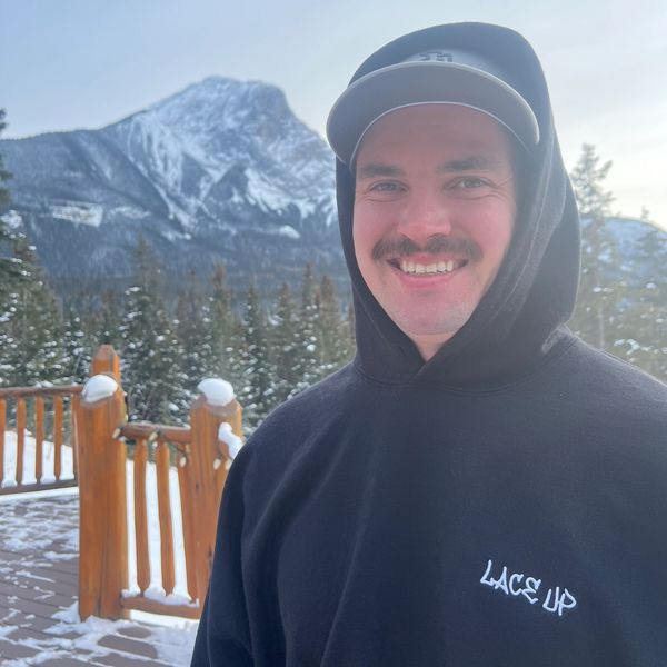 Smiling man in a black hoodie with snowy mountains in the background.