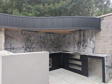 Modern outdoor kitchen with marble backsplash and black cabinetry under a shaded roof.