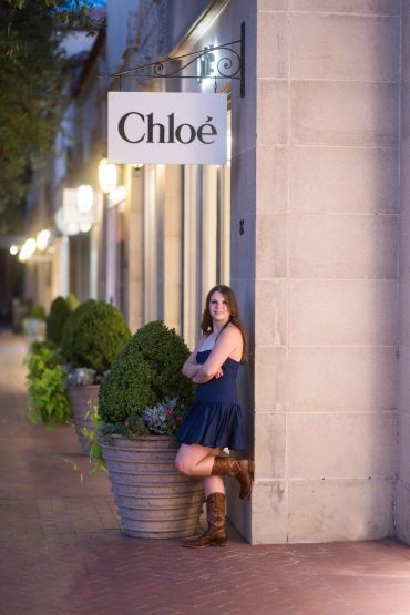 Young woman leaning against a wall outside a Chloe store at dusk.