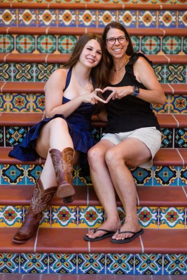 Two women sitting on colorful tiled stairs making a heart with their hands.