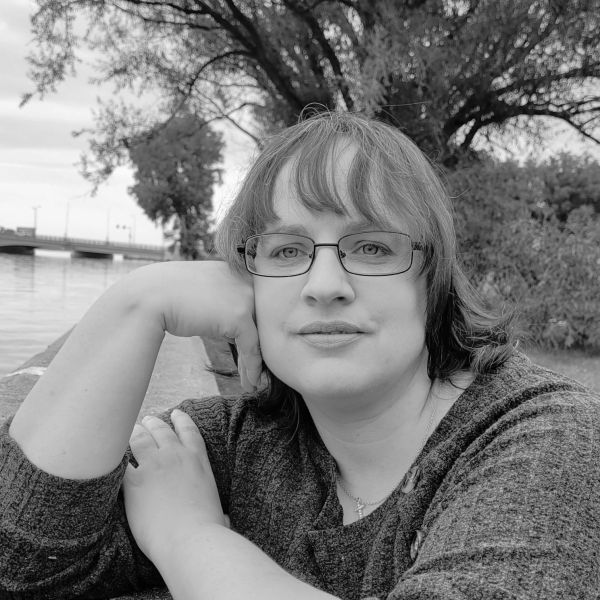 Woman with glasses posing by a riverbank in black and white.