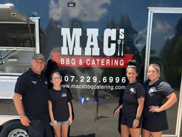 Four people stand in front of a MAC's BBQ &amp; Catering trailer.