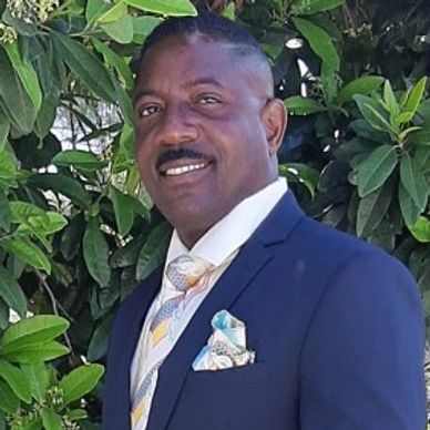 Man in a blue suit smiling outdoors with green foliage background.