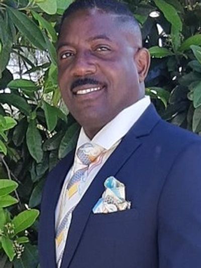 A man in a navy blue suit with a patterned tie and pocket square stands outdoors.
