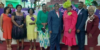 Group of eight adults dressed formally, posing indoors with purple decorations.