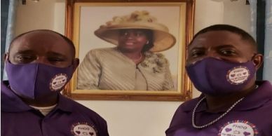 Two men wearing purple masks and shirts stand in front of a framed portrait of a woman.