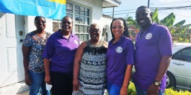Five smiling people standing outdoors in front of a house with a flag.