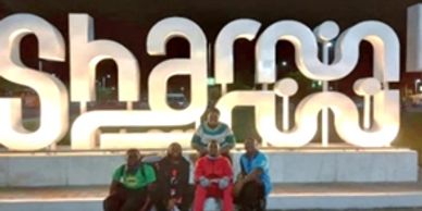 Five people posing in front of an illuminated Sharm sign at night.