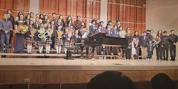 Group photo of children and adults on stage with a grand piano.