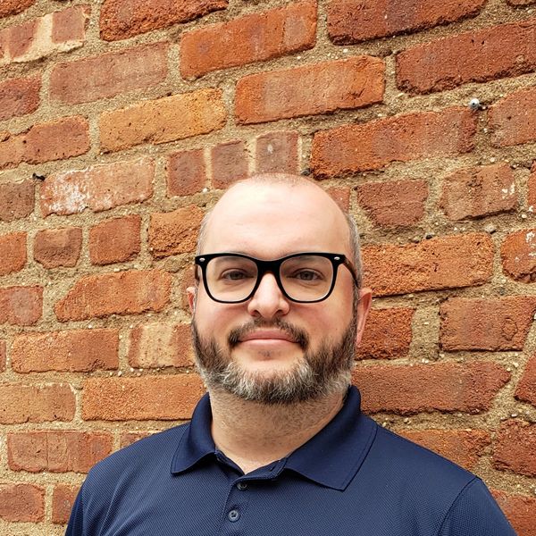 Man with glasses and beard posing against a brick wall.