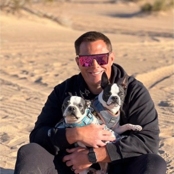 Man in sunglasses hugs two small dogs in a sandy area.