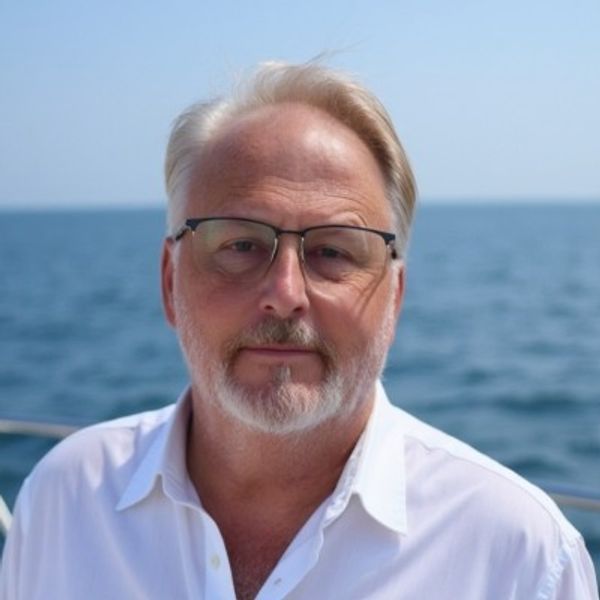 Older man with glasses and white beard on a boat with ocean background.