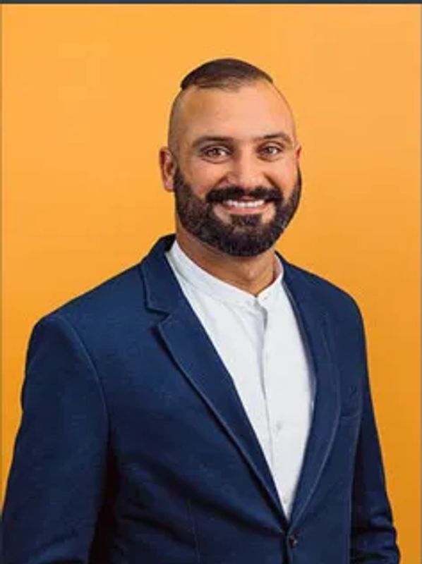 A smiling man in a navy blazer and white shirt against an orange background.