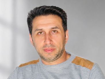 Portrait of a man with dark hair wearing a gray sweater with tan patches.