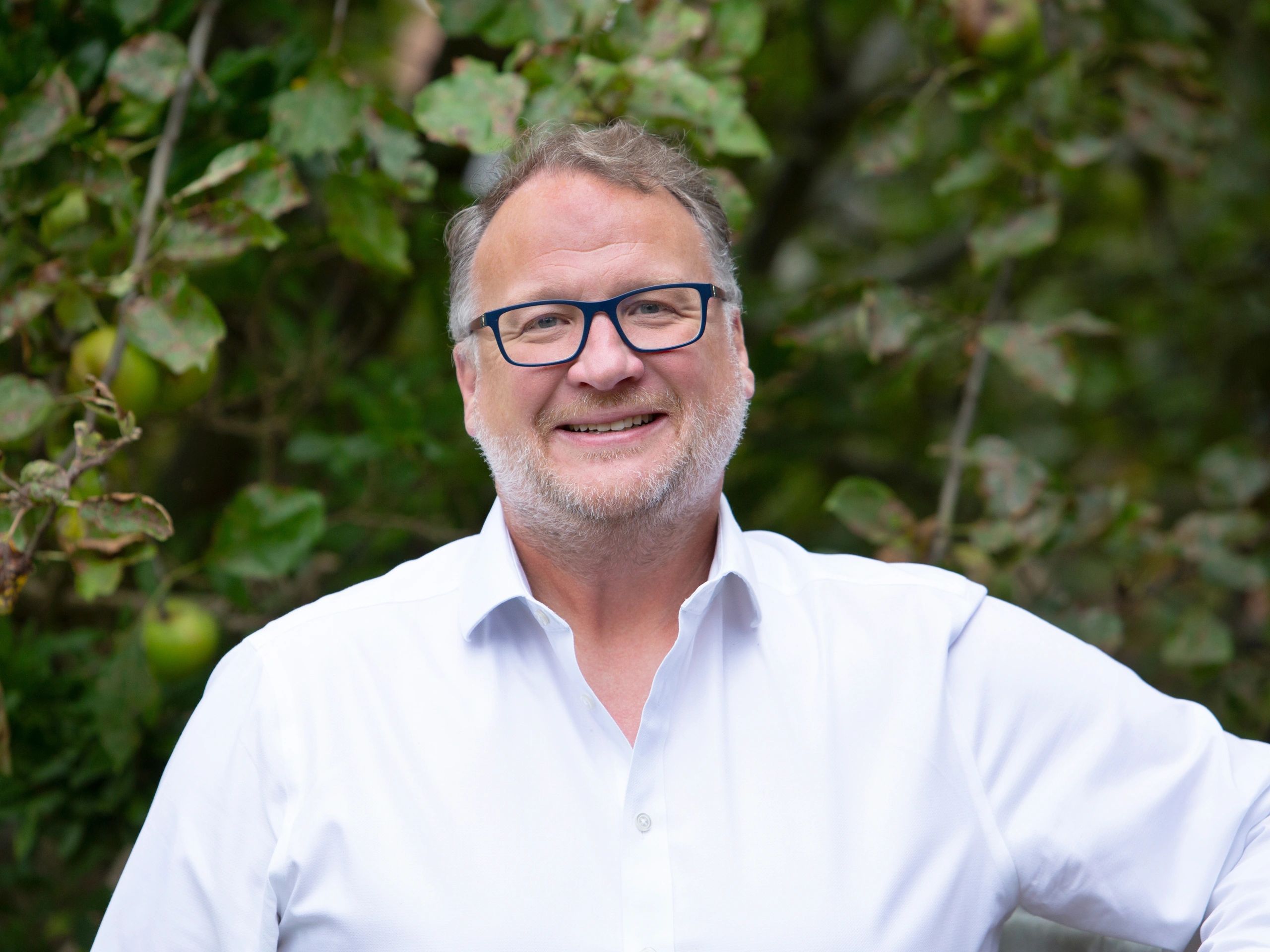 Smiling man with glasses in a white shirt outdoors with greenery.