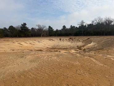 A large dry, cracked pond bed under a cloudy sky with trees in the background.