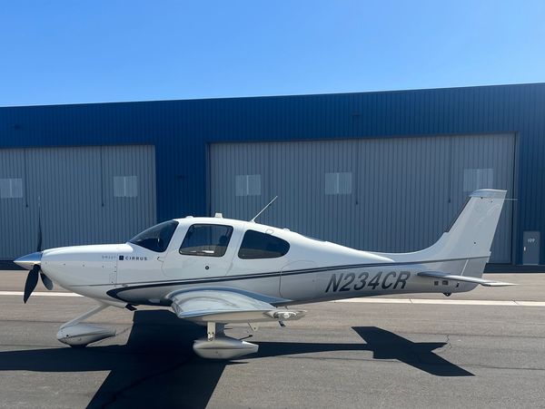 White Cirrus SR22T airplane parked on tarmac in front of hangars.