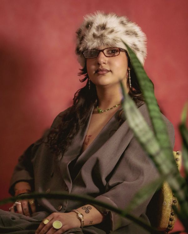 Person wearing a fur hat and glasses, posing confidently against a red backdrop.