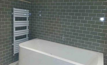Modern white bathtub against green tiled walls with a white towel radiator.