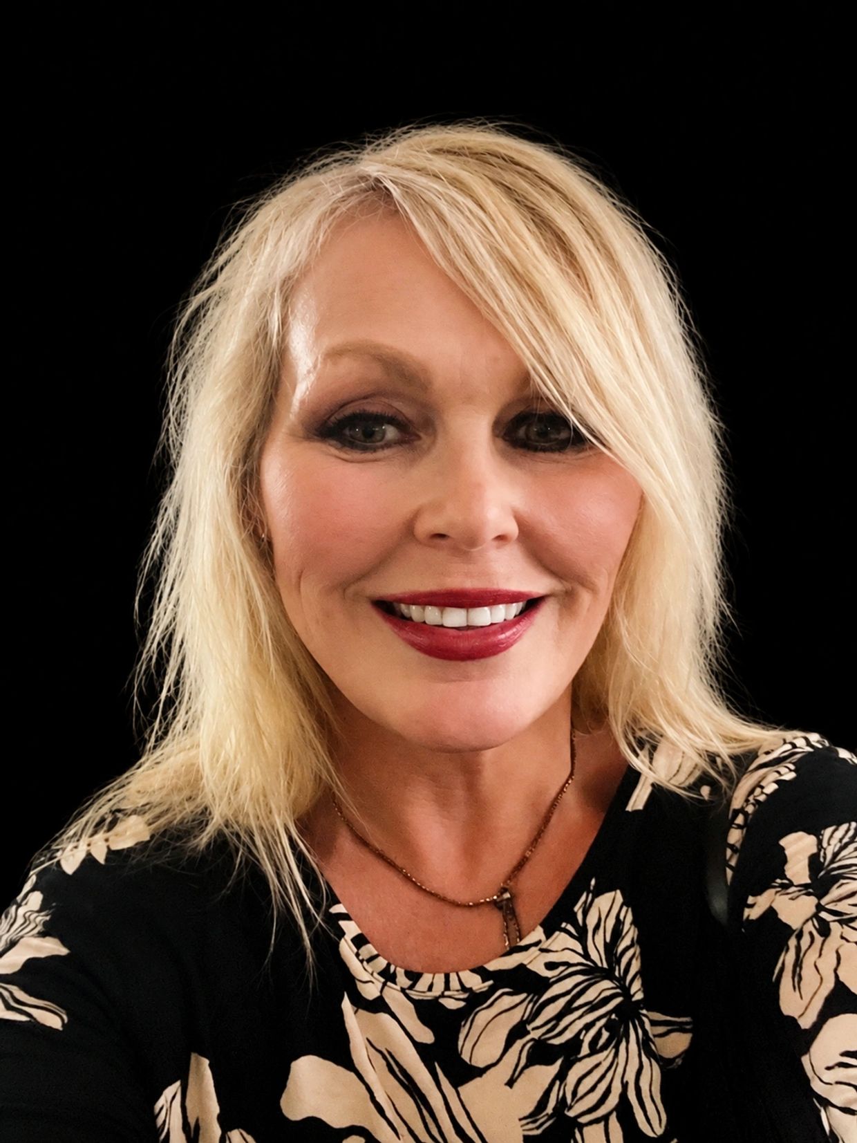 Smiling blonde woman with floral top on black background.