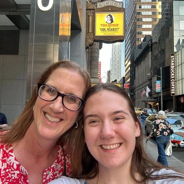 Two women smiling for a selfie on a bustling city street with theater signs.
