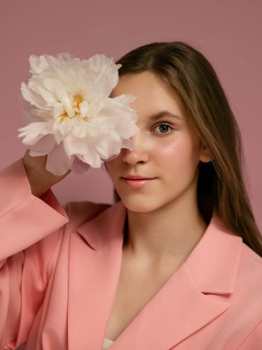 Young woman in pink blazer holding white flower over one eye.