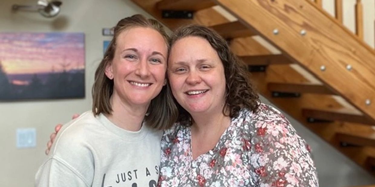 Two women smiling indoors by a wooden staircase.