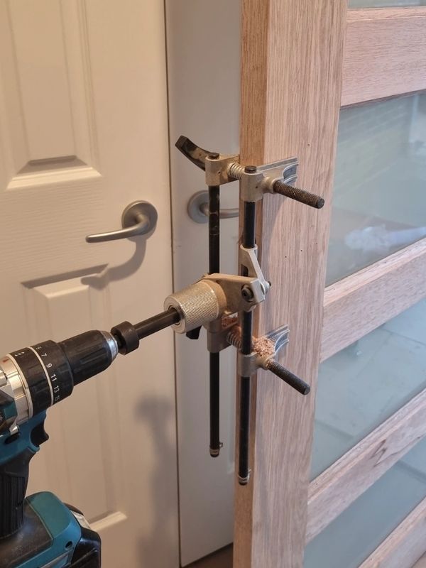 A drill with a hole saw bit attached to a wooden door clamp.