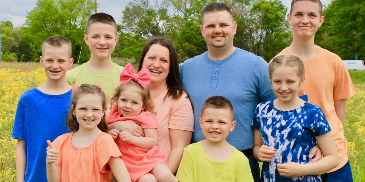 A happy family of ten posing outdoors in a field of yellow flowers.
