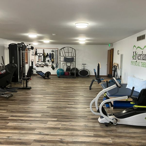 A spacious physical therapy gym with various exercise equipment and wooden flooring.