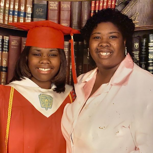 A proud graduate in red cap and gown smiles with a woman in front of a bookshelf.