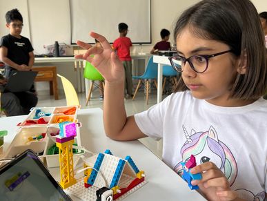 A girl builds a colorful LEGO structure while interacting with a tablet in a classroom.