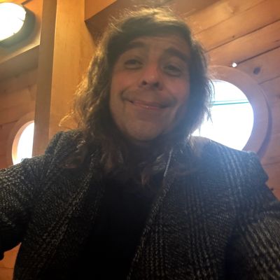 Person smiling indoors with wooden walls and round windows.