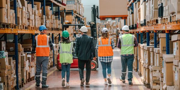 Team of workers, engineers, and manager walk warehouse aisle discussing operations and efficiency.