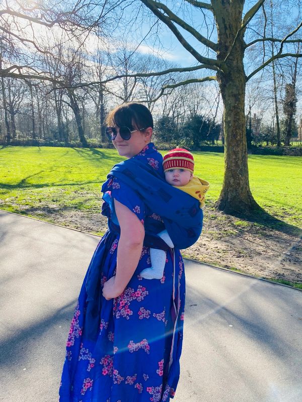 A caucasian woman in a purple dress carries her baby in a woven wrap sling.