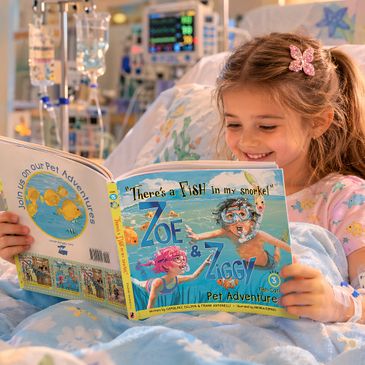A young girl in a hospital bed happily reading a colorful children's book.