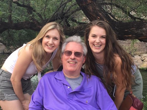 Two young women smiling with an older man outdoors under a tree.