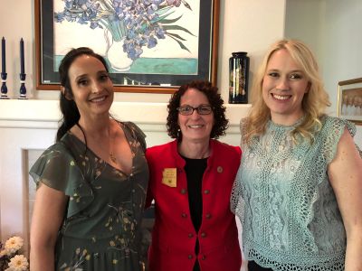 Three women smiling indoors, two in dresses and one in a red jacket.