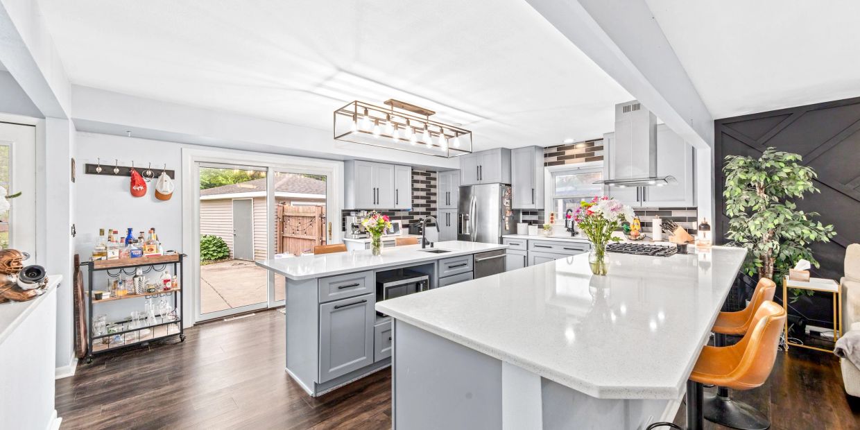 Modern kitchen with large white countertops, grey cabinets, and brown leather bar stools.