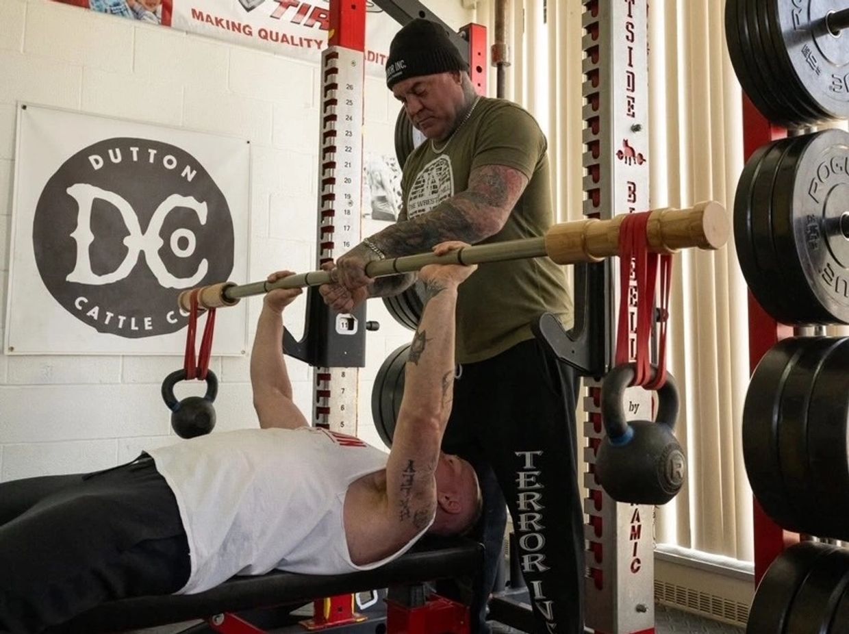 A man spots another man lifting weights with kettlebells attached.