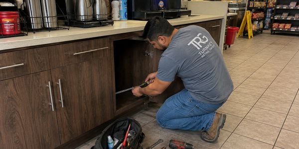 A technician repairs a cabinet in a convenience store, using tools from his bag.