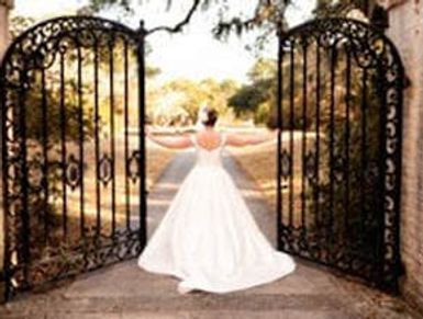Bride in white dress opening ornate iron gates on sunny day.
