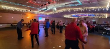 Couples dancing in a decorated hall with string lights and disco ball.